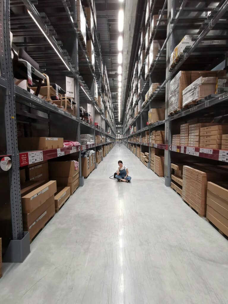 pexels-photo-2701434-2701434 An Asian woman sits in a large warehouse aisle filled with inventory racks and shelves.